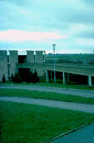 Photograph showing a raised concrete walkway running into the side of a building; behind, a view of hillside and distant houses can be seen; the photograph has been described as View of Town Centre, Peterlee