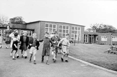 Photograph showing a group of six children, aged approximately seven years, walking towards the camera, down a drive leading from a low single-storey building with a central block with three large windows and wings on either side; a woman is walking behind the children; the building has been identified as Eden Hall Infants' School, Peterlee
