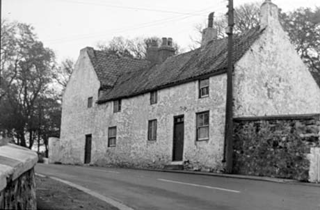 Photograph of the front and side of a roughly rendered cottage with three windows on the first floor and three windows and a narrow door on the ground floor; the cottage stands directly on the road and abuts a building at right angles or has a wing at right angles; the photograph has been identified as Old Shotton