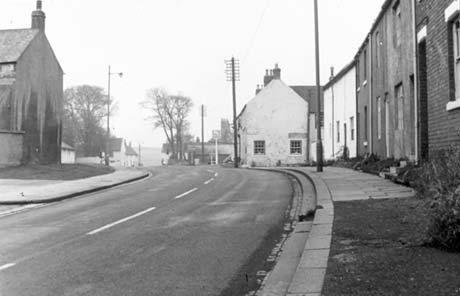 Photograph of a road running away from the camera; the end of a building and houses and fields in the distance can be seen on the left; on the right are terraced houses and the end of a building with an inn sign before it, which has been identified as the Black Bull; the photograph has been described as Old Shotton