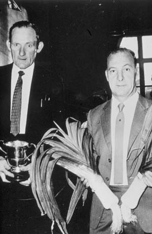 Photograph showing, on the left, a man in a suit and tie holding a trophy cup; on the right is a man in a suit and tie holding a large leek, who has been identified as J. Porter