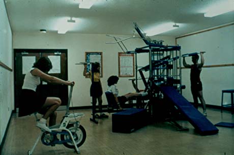 Photograph showing the interior of a small room with gymnastic equipment; a girl on the left is riding an exercise bicycle and the man and two women on the right are using a multi-gym; the photograph has been described as the Fitness Room in the Leisure Centre, Peterlee