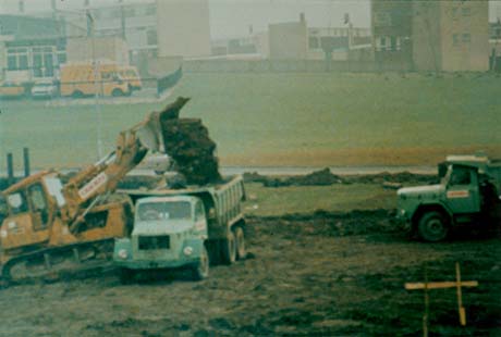 Photograph showing an earth-moving vehicle depositing soil in the back of a lorry; excavated ground can be seen in the foreground and buildings indistinctly in the background; the photograph has been described as Construction At Town Centre, Peterlee