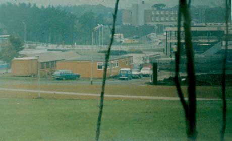 Photograph showing a low building with cars parked near it, a road beyond it and tall buildings beyond that; excavated earth can, possibly, be seen at the right of the photograph under a ramp; the photograph has been described as Construction At Town Centre, Peterlee