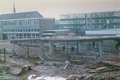 Photograph showing the roof of St. Cuthbert's Church above a building and another block of a building in the background; in the foreground, are debris on the ground and the skeleton of a ramp of concrete; the photograph has been described as Construction at Town Centre, Peterlee