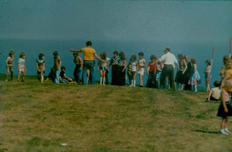 Photograph showing twenty four children, aged between seven and fourteen years, standing on the brow of a hill with sky only behind them; two men are attempting to organise the children, most of the children, and the men, have their back to the camera; more children can be seen on the right of the picture, which has been identified as British Legion Sports Day, Peterlee