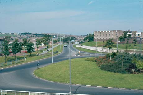 Photograph showing a roundabout in the foreground, with the road leading away from the camera; on either side of the road are buildings and there are houses in the distance; the photograph has been identified as Looking Along Essington Way, Peterlee