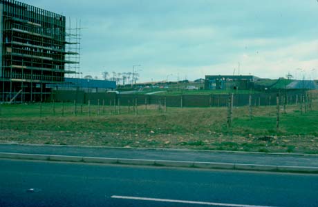 Photograph showing a road with a grass verges and a fence; beyond the fence are two low buildings; at the right of the picture is a tall building, of approximately ten storeys, surrounded by scaffolding; the photograph has been described as View of Factories, Peterlee