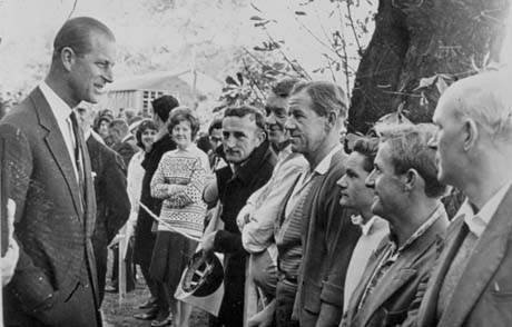 Photograph showing the Duke of Edinburgh, wearing a jacket and tie, on the left, speaking to approximately twenty people standing behind a barrier of string, with a tree and a hut behind them; they are informally dressed; the photograph has been identified as Visit By Duke Of Edinburgh, Peterlee