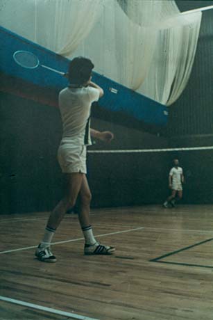 Photograph showing a man wearing shorts, shirt, socks and trainers, holding a raquet in his hand and with his back to the camera; he is standing on a court with a net and another man on the other side of the net; the photograph has been described as Playing Badminton At Leisure Centre, Peterlee