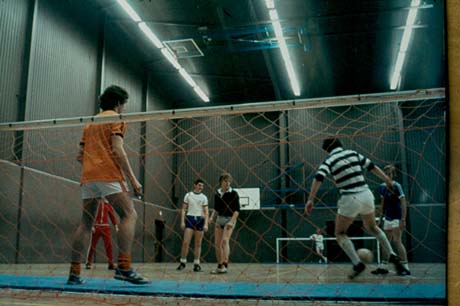 Photograph showing two boys, aged approximately fifteen years, wearing shorts with their backs to the camera, and five boys facing the camera, in a building of corrugated iron with fluorescent lighting; a net is in the foreground and a boy can be seen standing near a goal post; the photograph has been described as Playing Football At Leisure Centre, Peterlee