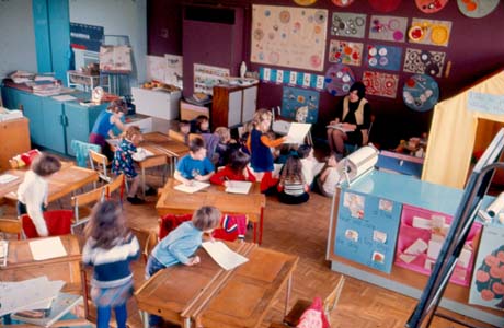 Photograph showing a classroom with ten desks, cupboards, drawings on the walls and approximately fifteen children, aged approximately four years, and a woman sitting on a chair near the wall of drawings; the photograph has been identified as the Nursery School at Peterlee