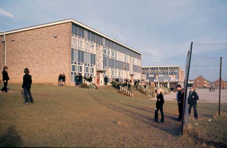 Photograph showing the exterior of a building with brick sides and a front of windows and blue panels at the top of a small slope; another block can be seen in the distance; approximately thirty children wearing blazers are standing in front of the building, which has been described as Grammar School, Peterlee