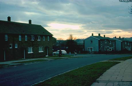Photograph showing a road running across the photograph with a block of brick houses and semi-detached houses on the opposite side; there are grass verges on either side of the road; a car can be seen parked in a side road and figures can be seen near the car; the photograph has been identified as Yoden Road, Peterlee