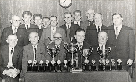 Photograph of a group of fifteen men posed against the wall of a room with a table in front of them containing four trophy cups, a trophy on a stand and thirteen individual small trophies; the men are dressed in suits and ties, and have been identified as follows: Back Row, left to right: Tom Stubbs; Bill Harrison; Alfie Thompson; Jack Wilkinson; Will Charlton; Jim Wilkinson; Simon Richardson; [ ]; Don Wright; Danny O'Connor; Front Row, left to right : [ ]; Ned Robinson; Harry Adams; Tom Barron; Jack Nicholson; they have been identified as trophy winners at Seaham conservative Club