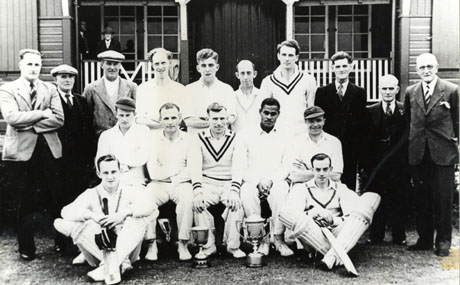 Photograph of eleven men in cricket whites posed in front of a pavilion six men in suits; behind them two men can be seen on the veranda of the pavilion; in front of the group there are two trophy cups on the ground; they have been identified as Seaham Park Cricket Team, as follows : Middle Row second left : Arthur Lanes; Back Row, third right : Tom Finkel; Mr. Adams; Harry Wakefield, bottom right