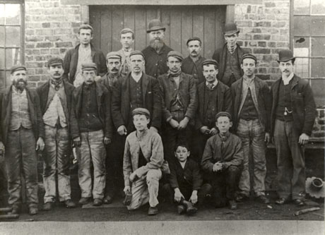 Photograph of a group of eleven men in work clothes of waistcoat, trousers, jacket, scarf, and cap, accompanied by three men in suits, ties and bowler hats, and by three boys, posed outside a brick building, possibly a colliery building; they have been identified as joiners and fitters from New Seaham Colliery