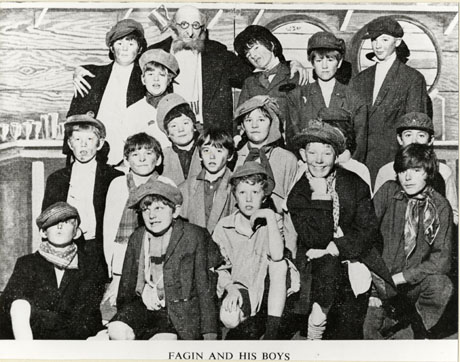 Photograph of a group of seventeen boys, aged approximately twelve years, in caps, jackets and scarves, posed with a man wearing a false beard, an overcoat and a scarf, against scenery depicting barrels, glasses, and wooden walls; they have been identified as part of the cast of the production of Oliver produced by North Lea School