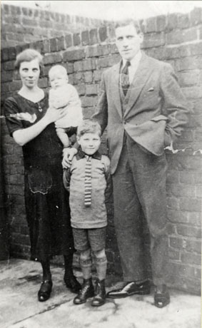 Photograph of a woman dressed in a dark dress, holding an infant, standing with a man dressed in a suit and tie, and with a boy of approximately seven years in front of a brick wall; the boy is wearing boots, shorts, a jumper and a striped tie; they have been identified as Mr. and Mrs. Dick and Family, Seaham