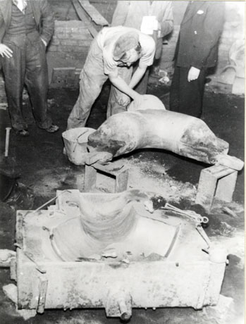 Photograph of a man in work clothes bending over a curved object which is balanced on two boxes; behind him the legs and torsoes of three men can be seen; at the front of the picture is an oblong container in which a depression has been made; the picture has been described as a template used in the making of a sand-mould at Seaham Foundry, in approximately 1950s