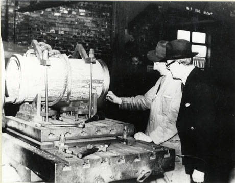 Photograph of a man in an overall and a Trilby hat, standing with a man in a suit and Trilby hat, looking at a cylindrical piece of machinery on a bench; behind them is the wall of a workshop; the photograph has been described as casting out of mould at Seaham Foundry in approximately the 1950s
