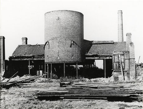Photograph of single storey building of brick with slates on its roof and without walls other than at its end; in the middle of the building is a large round drum-like structure rising high above the roof of the building; four chimneys rising from the ground above the roof-line can be seen at the corners of the building; in front of the building are piles of timber on the ground; the building has been identified as the Retort House at Seaham