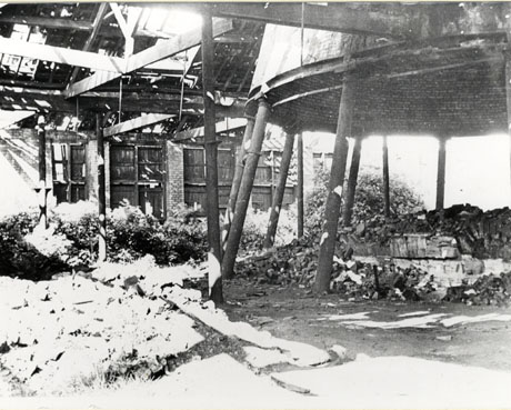 Photograph showing steel pillars supporting a roof of girders seen from below; to the right of the picture are the pillars supporting a circular brick structure which can be seen from below and the remains of a hearth below it can also be seen; behind the pillars a brick wall with large wooden doors in it can be seen; it has been described as Inside of Retort House, Seaham