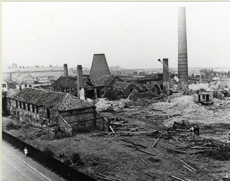 Photograph of Seaham Bottle Works seen from above; on the left of the picture is a rectangular building of brick, in a ruinous state, seen from its end and side; at right angles to it is large building, with a kiln-like structure in its middle and three square chimneys, all also in a ruinous state; to the right of the picture are two round chimneys; the whole site appears to be in the course of demolition and a mobile crane can be seen on the right of the picture; the site is that of the bottle works at Seaham