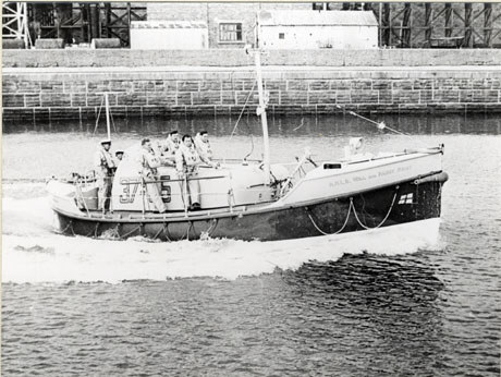 Photograph of a lifeboat in a harbour; the boat is moving from left to right of the picture in front of the wall of a harbour; six men are on the boat wearing life jackets; the number of the boat is 37-[ ]5 and its name is Her Majesty's Life Boat Will and Fanny K[esy]; the boat has been identified as being launched in Seaham