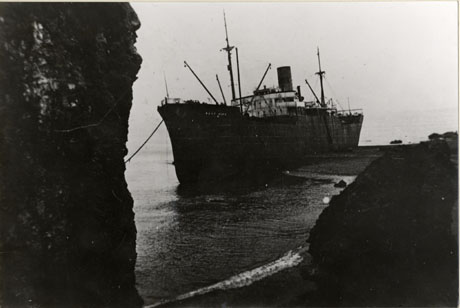 Photograph of a steam ship, identified as the West Hikaaground near Seaham, showing a ship with one funnel in shallow water near the shore; the side and the superstructure of the ship can be seen; a cliff and rock are in the foreground of the picture