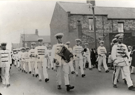 Photograph of three lines of young men in light uniforms with dark frogging, walking towards the camera, past the ends of terraced houses; further members of the parade can be seen in the distance beyond the young men; the parade has been identified as Dawdon Carnival