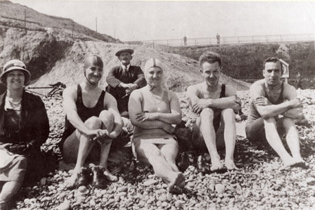 Photograph of two men and two women in swimming costumes sitting on a beach of pebbles with dunes, and a harbour wall to the right, behind them; a middle-aged woman, fully clothed, is sitting to the left of the swimmers and a fully clothed middle-aged man is sitting behind them; a bicycle is lying on the ground behind the group; they have been identified as the Sandell family at Seaham