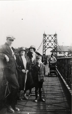 Photograph of a young man, wearing a suit and cap, leaning on the parapet of a bridge; two middle-aged women, wearing coats and cloche hats, and a boy, wearing a long jacket and shorts, are standing near him; a woman pushing a perambulator is crossing the bridge towards the camera; the surface of the bridge and the piers from which it is suspended can be seen; it has been identified as Dawdon Suspension Bridge