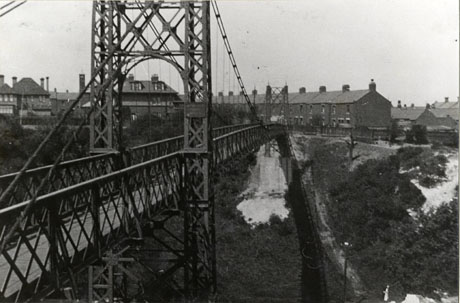 Photograph of the piers and roadway of a suspension bridge with the bridge running away from the camera; beyond the bridge, a row of terraced houses and large detached houses can be seen; the photograph has been described as Dawdon Suspension Bridge, Green Drive