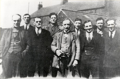 Photograph of nine men posed at the rear of an outhouse attached to a larger house; they are wearing suits or overcoats; they have been identified as members of New Seaham Conservative Club