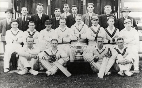 Photograph of twelve men in cricket whites posed with nine other men, in suits, outside the pavilion; between the players on the front row is a trophy cup; two of the players on the front row are wearing cricket pads and holding cricket bats; they have been identified as members of Seaham Park Cricket Team