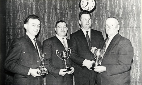 Photograph of four men, all wearing suits, posed against a wall inside a building; three are holding trophies and the fourth is shaking hands with the man on the extreme right; they have been identified as winners of the Bowls League at seaham Conservative Club