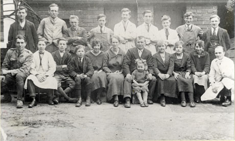 Photograph of two men wearing striped aprons, four men wearing white coats, one man wearing overalls, one man wearing a military style jacket, four men wearing suits, five women, three boys and a small girl posed in front of a brick building; they have been identified as New Seaham Store Staff