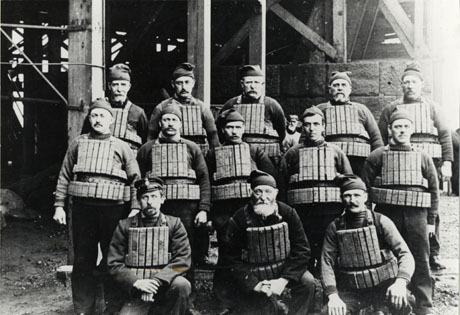 Photograph of thirteen men wearing lifebelts, made of what appears to be cork, and woollen hats, apart from the man at the left of the picture who is wearing a peaked cap; they are posed under the legs of a bridge or jetty; they have been identified as the crew of the lifeboat Skyer