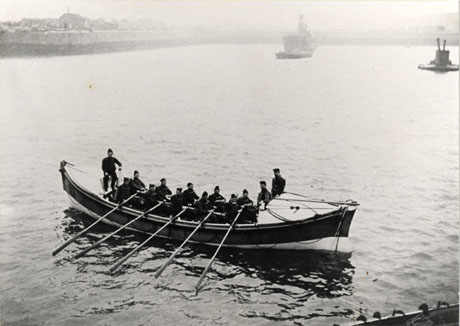 Photograph of a boat, in the foreground, being rowed by ten men, who are being observed by three other men ; in the distance, a large ship can be seen proceeding towards the camera; also in the distance, the walls of a harbour can be seen; the photograph has been described as Seaham Lifeboat Crew