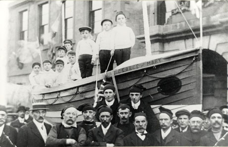 Photograph showing a lifeboat with the name George and Mary, in front of a stone building with sash windows and an arch; nine small boys are standing in the boat; ten members of the lifeboat service can be seen, along with crowds, below the boat; the boat has been identified as being in Seaham