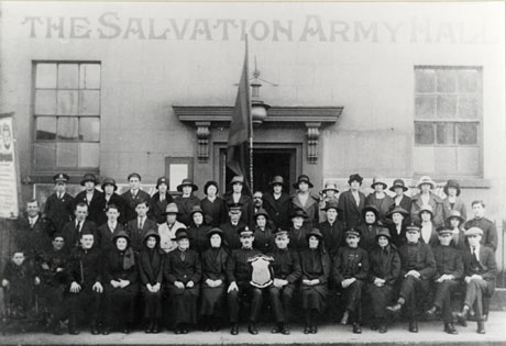 Photograph of the exterior of The Salvation Army Hall, Seaham, showing a stone building with long sash windows and an imposing doorway; approximately forty four people, some of whom are in Salvation Army uniform, are posed in front of the hall; a banner can be seen on the left of the picture and a flag in the centre; a trophy is being held by a man on the front row; a child at the extreme left appears to be in a wheel chair