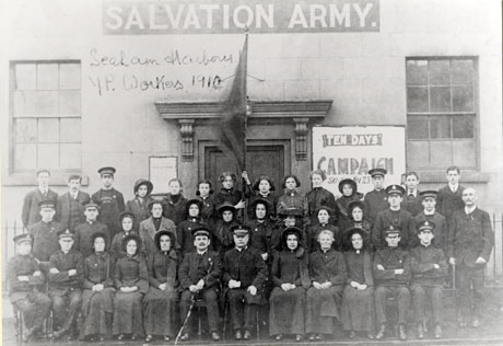 Photograph of the exterior of the Salvation Army Hall at Seaham, showing a stone building with sash windows and an imposing doorway; a group of thirty eight people, twenty two of whom are female, is posed outside the building; a poster reading Ten Days Campaign Sat [ ] Nov 23.... can be partially seen behind the group; the words Seaham Harbour Y. P. Workers 1910 has been written on the photograph