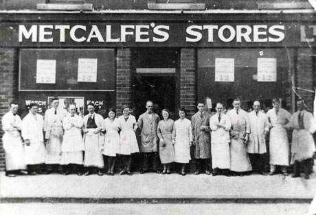 Photograph of twelve men and four women posed outside Metcalfe' Stores Ltd., Seaham; nine of the men are wearing long aprons and three are wearing overalls; the women are all wearing overalls; the windows of the shop can be seen, but only notices announcing the price of goods can be discerned
