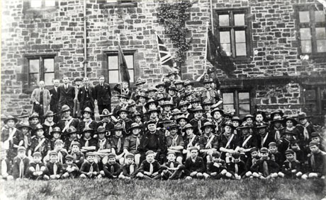 Photograph of a group of approximately seventy scouts and cub scouts, posed in front of a stone building with mullioned windows; on the left at the back of the group are six men in suits and ties; in the middle of the group is a man dressed as a clergyman; three flags can be seen at the back of the group; the group has been described as Seaham Colliery Scouts, Easter Sunday, 1934