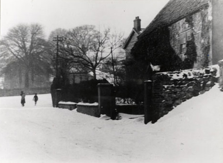 Photograph showing, on the right of the picture, part of the exterior of a building covered in ivy; in front are the walls of the garden; in the distance is an indistinct building which may be a church, surrounded by trees; the foreground of the picture is the surface of the road covered in snow; two indistinct figures can be seen walking on the road; the building has been identified as Ivy Cottage, Dalton le Dale