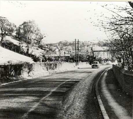 Photograph showing a road curving away to the right away from the camera towards a building with a hillside beyond it; a slope behind a fence is on the left of the picture; on the right, part of a wall can be seen ; a car is travelling along the road towards the camera and another can be seen in the distance