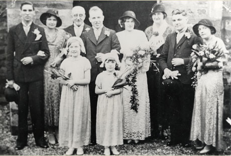 Photograph of four men and four women standing in a group outside a stone wall; the men are wearing suits and button holes; the women are wearing long dresses and hats and are carrying bouquets; two girls of approximately six and ten years are standing in front of the group wearing long dresses and carrying bouquets; they are described as Wedding Group, Seaton