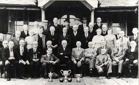 Photograph of a group of twenty nine men and four women posed outside a low building with a veranda; most of the group are elderly and are wearing suits; in front of the group are three trophy cups and two bowling balls; they have been identified as members of Seaham Park Bowls Club
