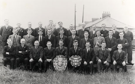 Photograph of thirty one men, wearing suits and ties, posed on grass with the end of a house in the distance; in front of them are two trophy shields; they have been identified as members of the Seaham Conservative Club Committee and members of the Club, photographed on the occasion of their winning the Tyne and Wear Trophy